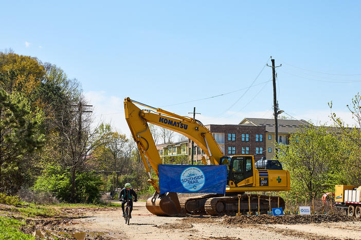 A large construction machine with a crane arm holds a sign saying "southside trail" over a dirt path.