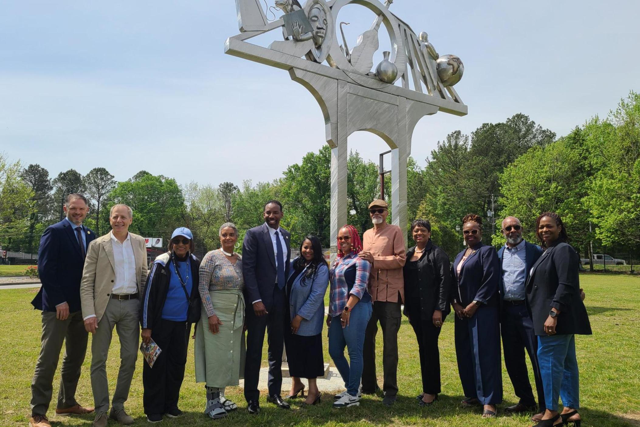 the mayor and a group of artists stand shoulder to shoulder in the grass in front of a large metal monument. It has an arch with a flat top where music objects, a globe, and the face of Martin Luther King are visible on top.