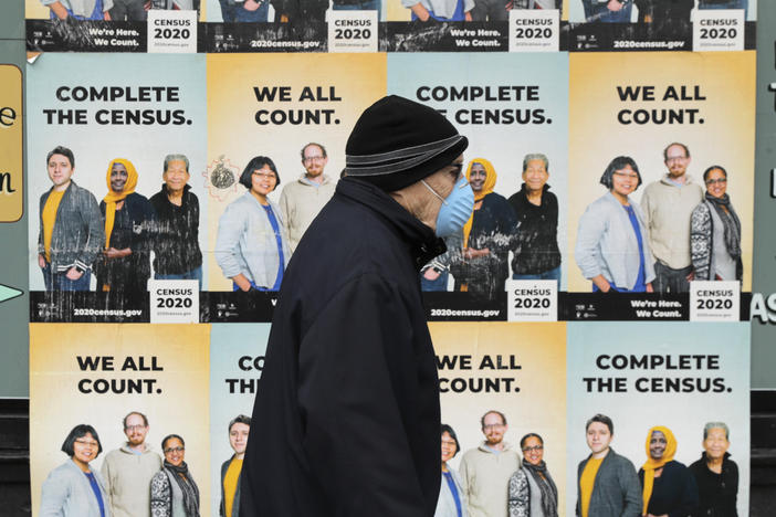 In this April 1, 2020, file photo, a man wearing a mask walks past posters encouraging participation in the 2020 Census in Seattle's Capitol Hill neighborhood. A delay in census data is scrambling plans in some states to redraw districts for the U.S. House and state legislatures. The Census Bureau has said redistricting data that was supposed to be provided to states by the end of March won't be ready until August or September. (AP Photo/Ted S. Warren, File)