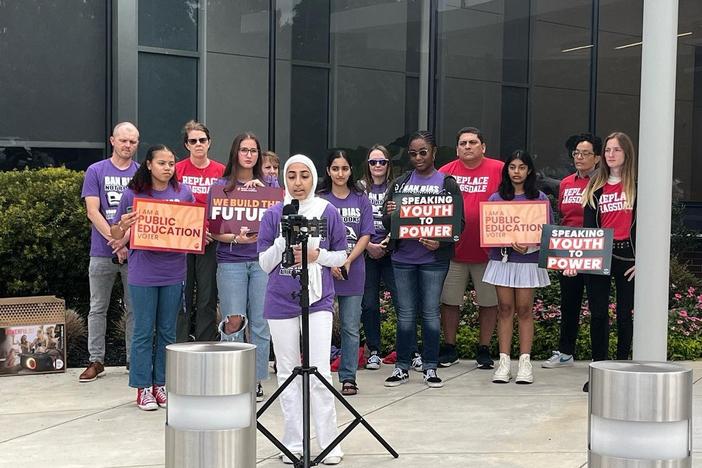 Georgia Youth Justice Coalition representative Haya Fatmi, wearing a white hijab and purple shirt that says "ban bias not books", speaks at a press conference before the Cobb County Board of Education meeting September 15, 2023. More students stand behind her in matching shirts holding signs. 