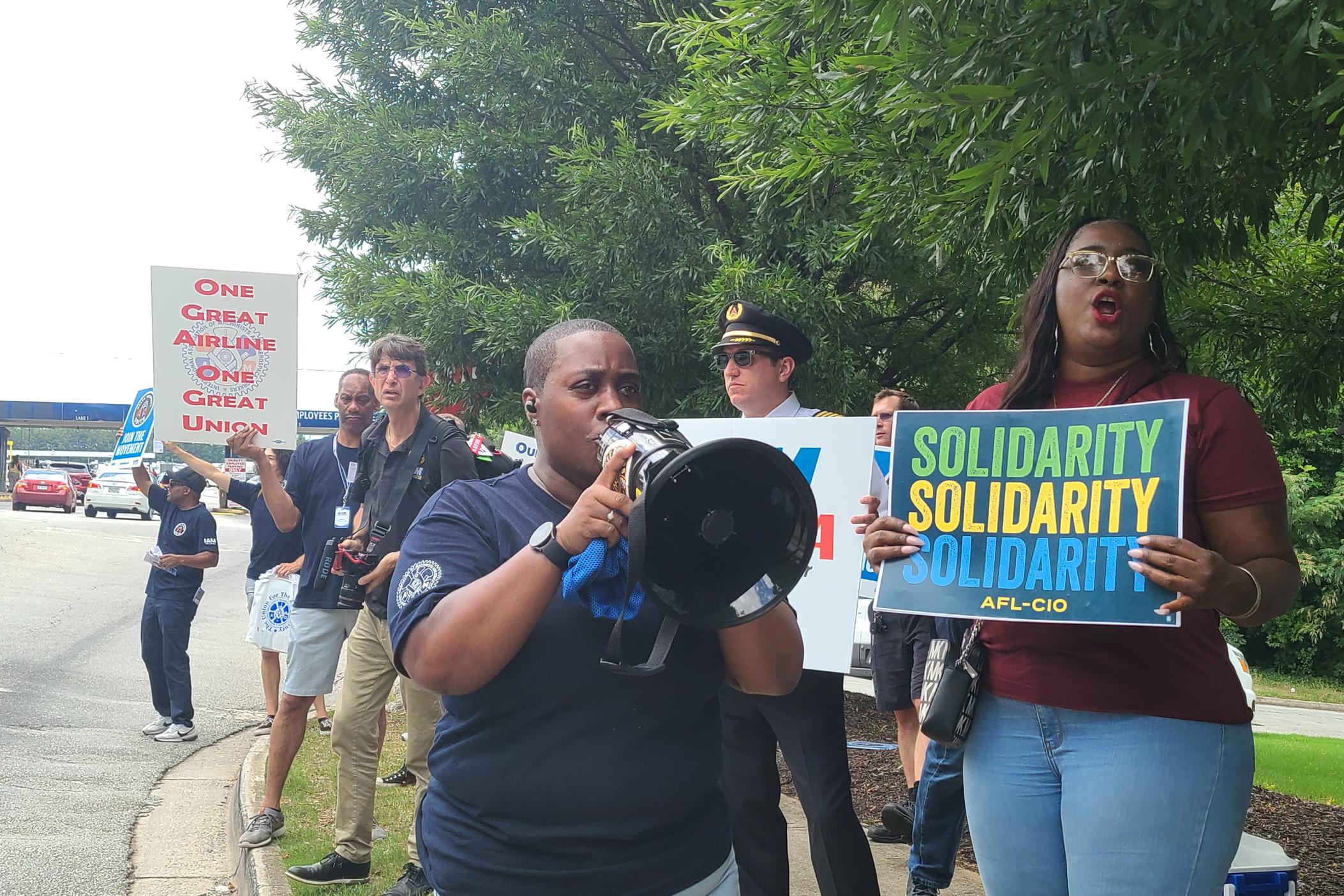 Avery hold a megaphone wearing a black shirt and several people stand behind her with signs saying " solidarity" and "one great airline, one great union"