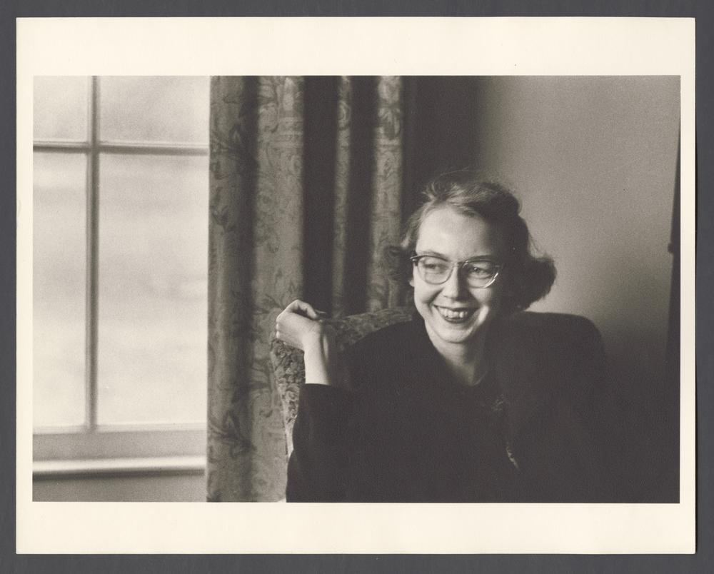 Flannery O’Connor, smiling, sitting by a window.