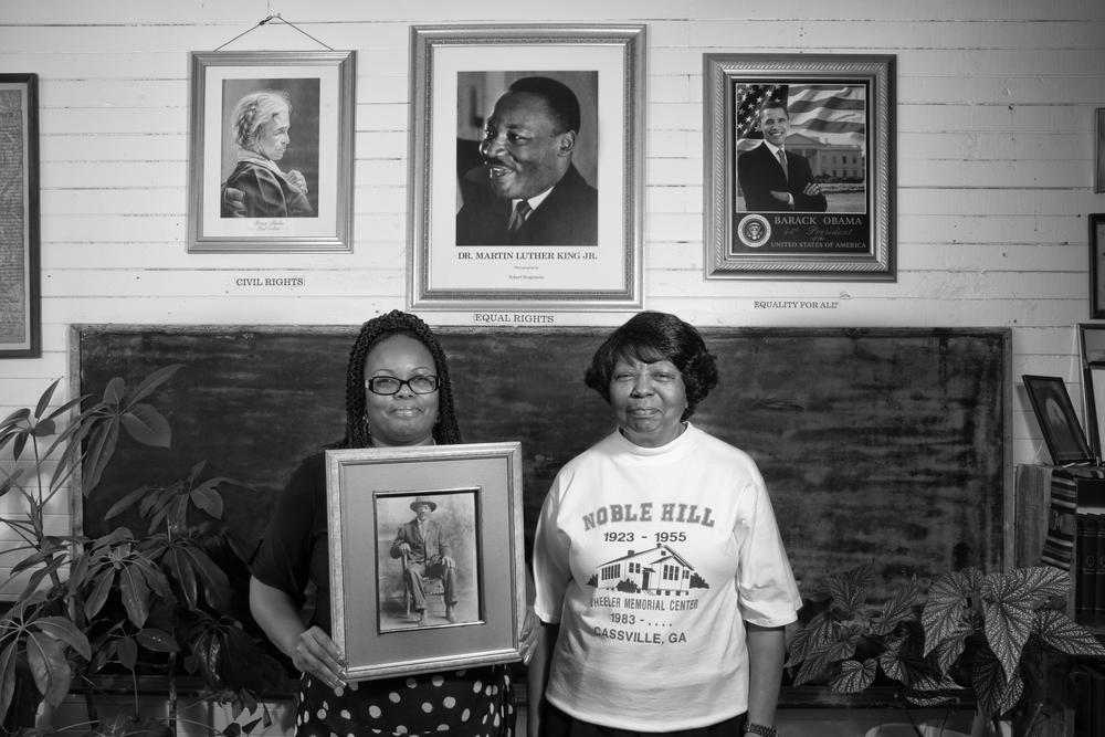 Two women stand in a Rosenwald school holding history artifacts from the school's history.