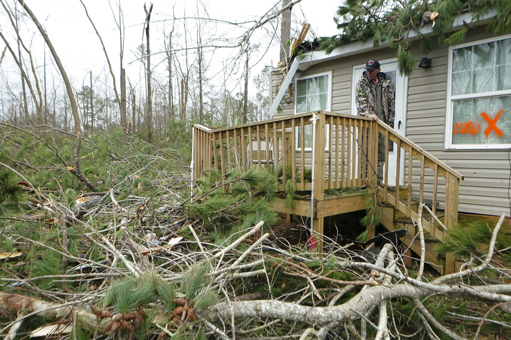 A man stands on the wooden porch of a home, looking at a tree that has fallen on his home.
