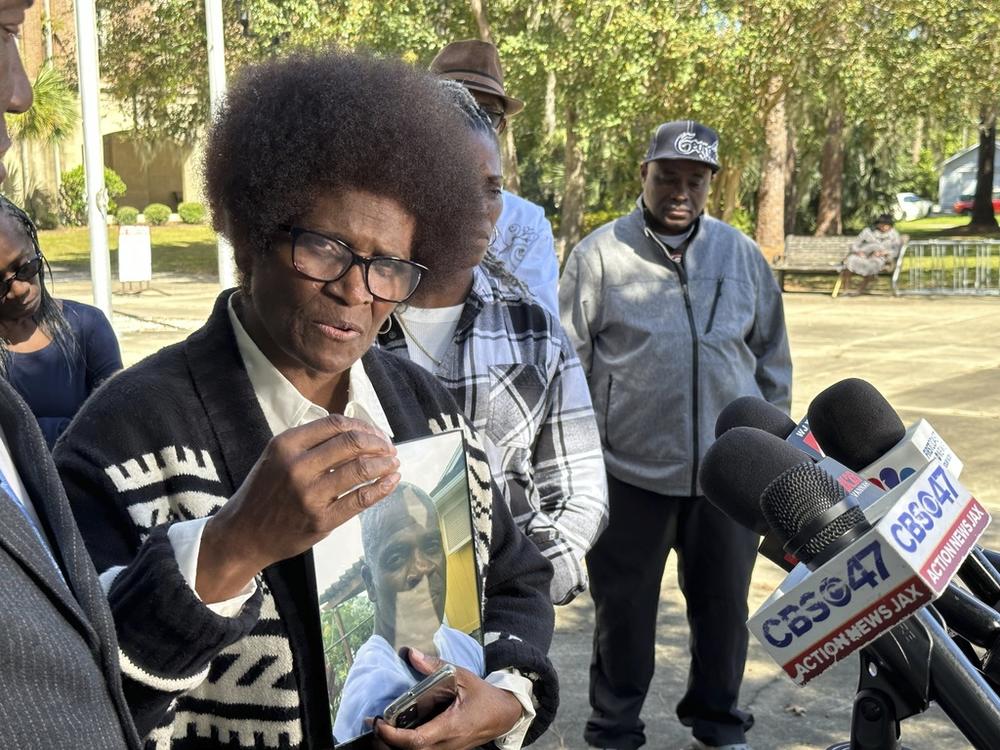 Mary Cure is shown holding a picture of her son Leonard Cure as she speaks in front of microphones,