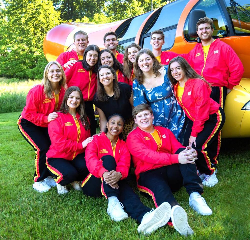 Out of more than 7,000 applicants for the 2024-25 season, Oscar Mayer selected these 12 Wienermobile drivers, posing with Wienermobile coordinator Emily Schmitt (blue dress) and head dog Molle Twing (black dress) in Madison, Wisconsin, where the drivers trained for two weeks in June 2024. Courtesy of Sarah Oney