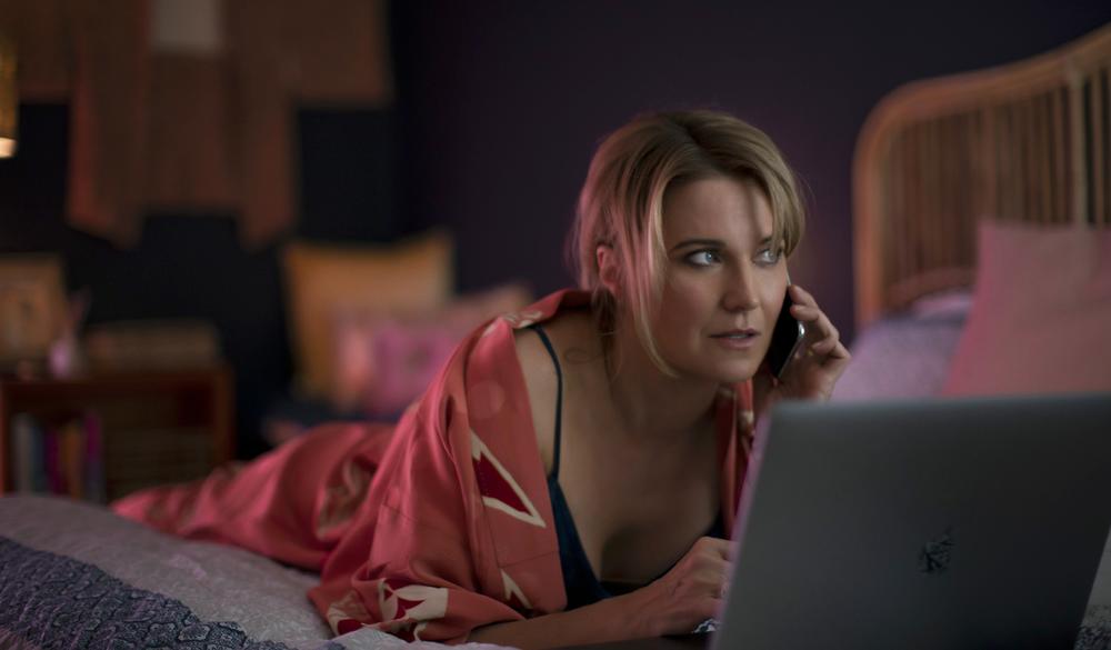  A woman lying on a bed talking on a cell phone looking into a laptop screen.