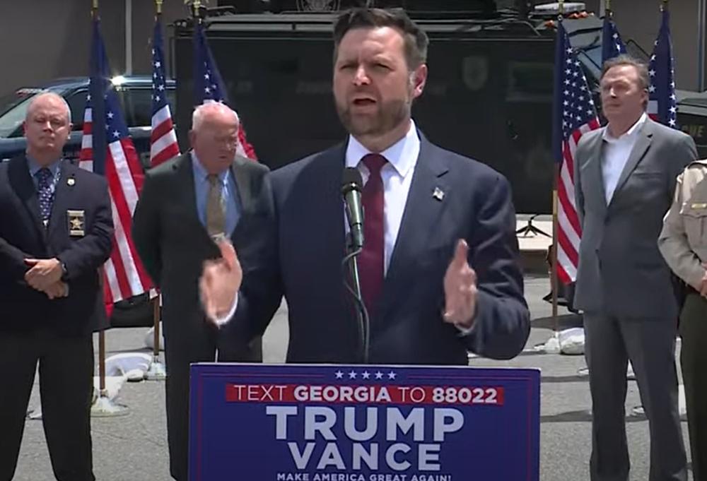 Ohio Sen. J.D. Vance is shown speaking from behind a podium.  He is flanked by other men and flags.