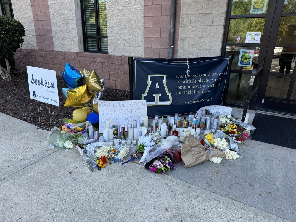 Flowers, balloons, signs, candles and other symbols of support are shown on a sidewalk next to a door.