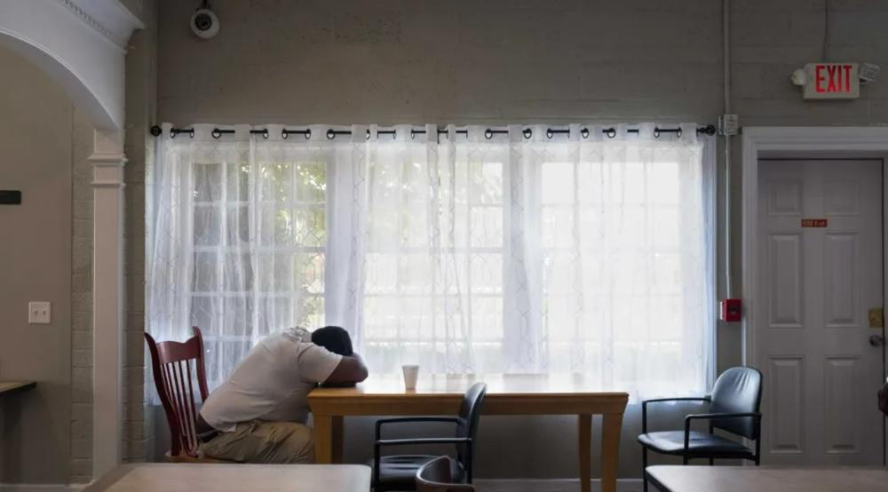 A man is shown sitting on a chair with his head facing down onto his arms resting on a table inside an empty room.