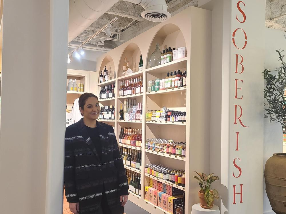 Soberish owner Mehrnush Saadat leans in a dark jacket against a wall with the word "soberish" painted vertically on the wall next to her.