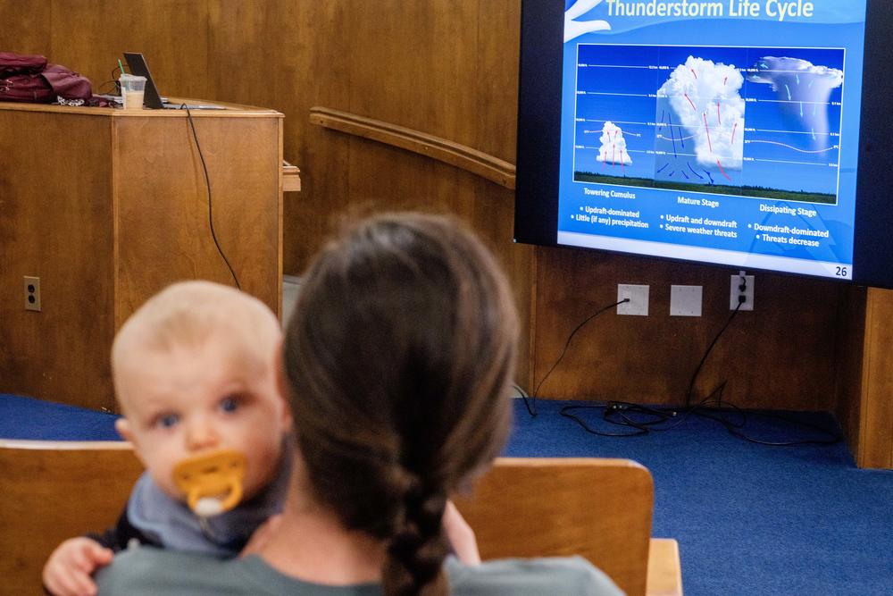 A mother with an infant on her shoulder tries to view a screen showing the three stages of a storm lifecycle during training to become a National Weather Service trained weather spotter. 