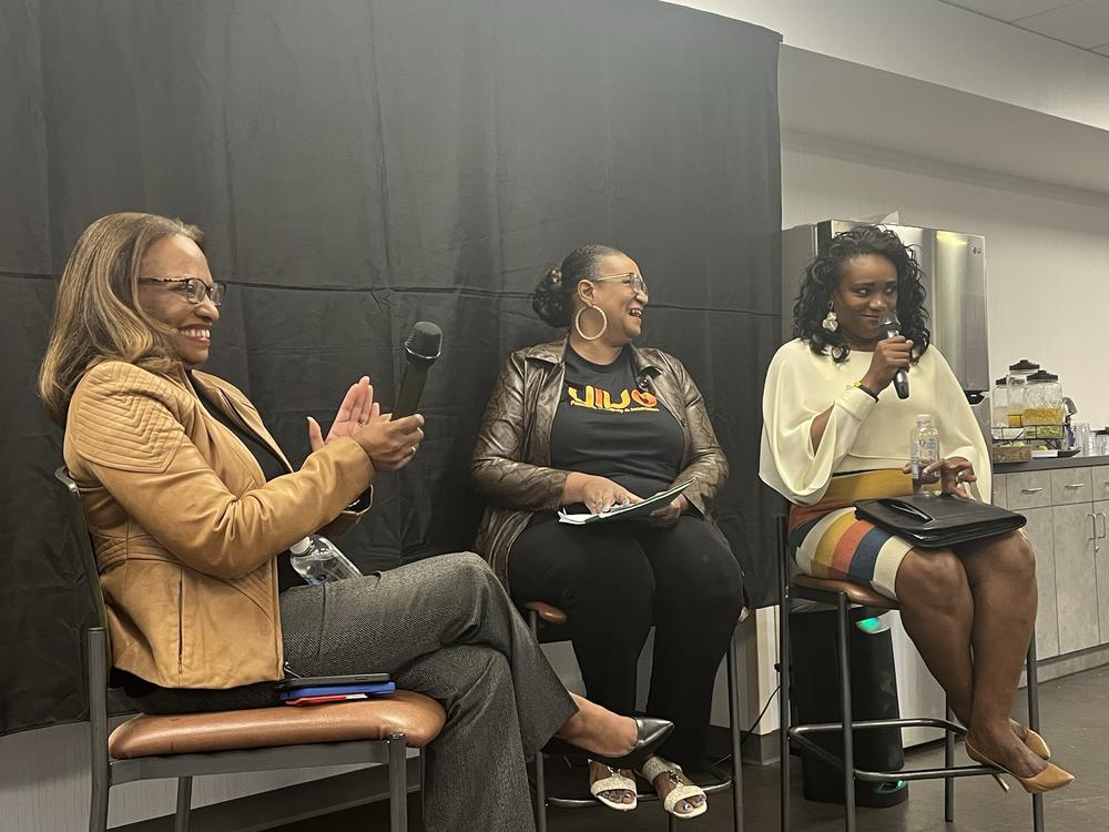 Acting Secretary Todman sits on a chair against a black background with two other black women of Caribbean descent in Norcross.