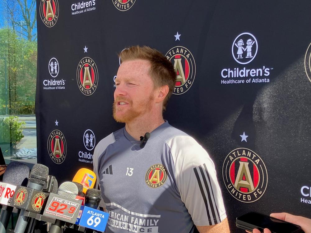 Dax McCarty is shown in an Atlanta United uniform speaking behind a podium.