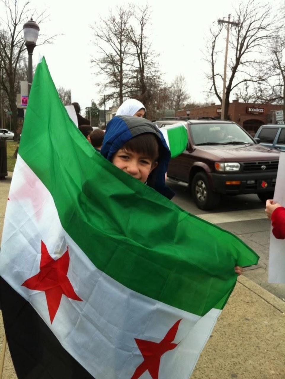 A child is shown in a parking lot holding a very large Syrian flag.