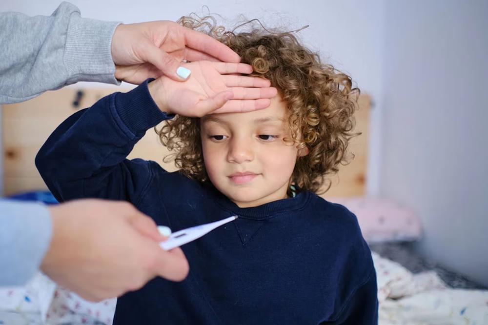 A child holds their hand to their forehead while an adult holds a thermometer.