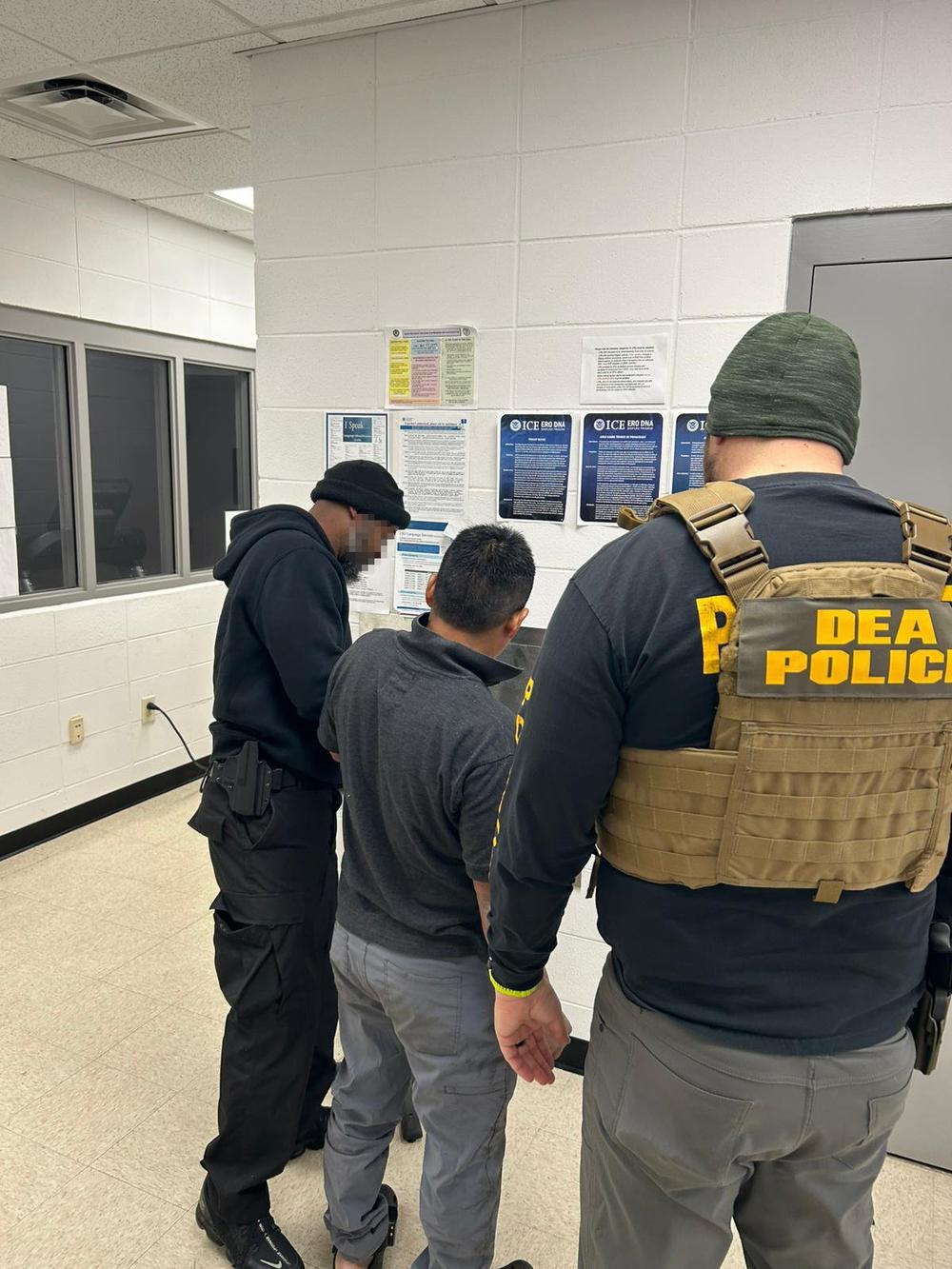 Three individuals are shown from behind, standing in a drab room.  One of them wears a uniform that says "DEA Police."