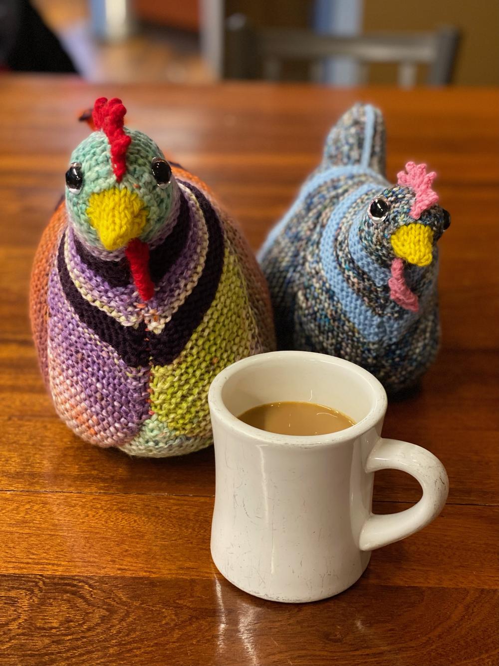 Two knitted chicken toys made out of multicolored yarn posed beside a coffee mug