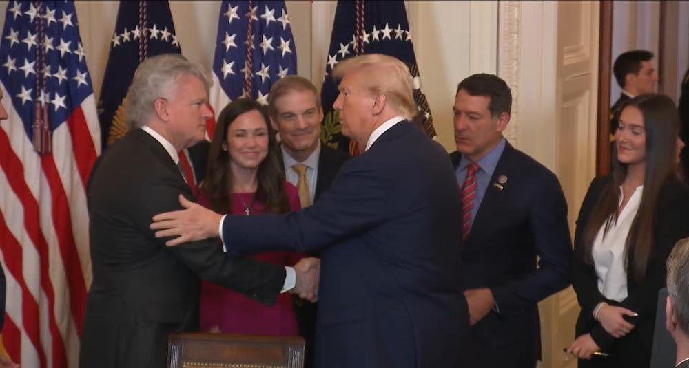 President Donald Trump is shown shaking the hand of Rep. Mike Collins while the family of Laken Riley and others look on, in a White House setting surrounded by American flags.