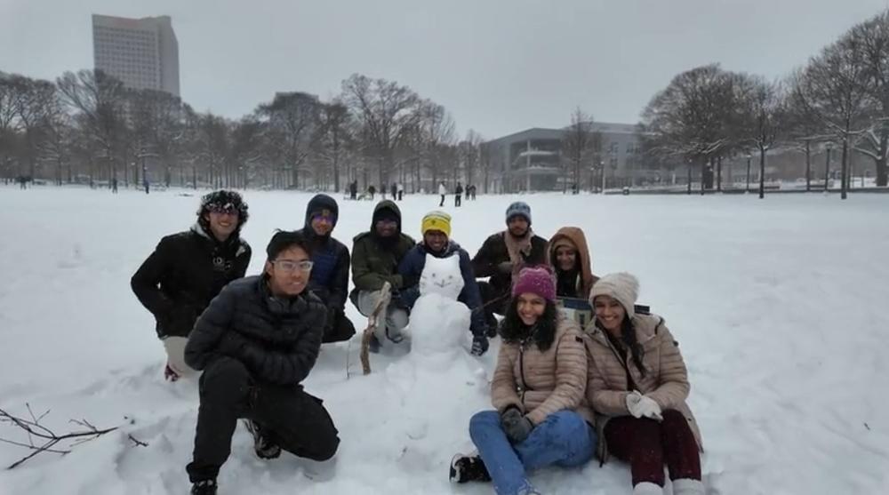 Nine students in winter clothing are shown outside in a snowy field seated on the ground near a snowman.