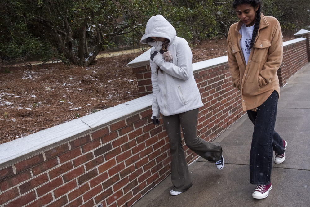 Two people are shown walking on a sidewalk as snow lightly falls on a wall.