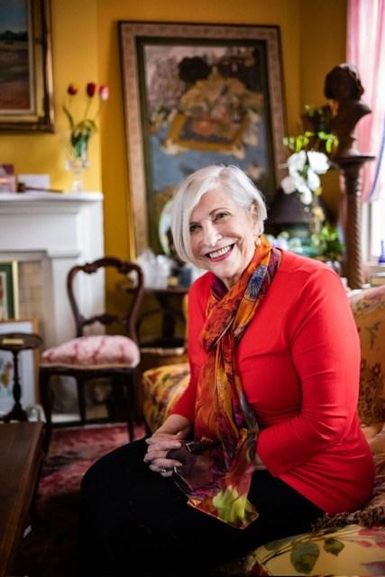 Nathalie Dupree is shown smiling and seated inside a floral and colorful domestic setting.