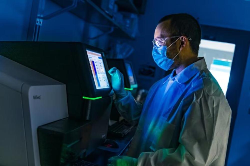 A man in a lab coat and medical mask peers at a lit medical monitor in a darkened room.