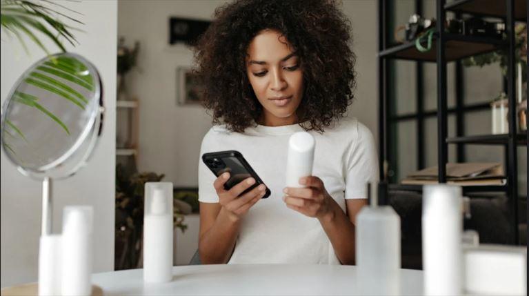 A Black woman looks at a beauty product and holds a cellphone