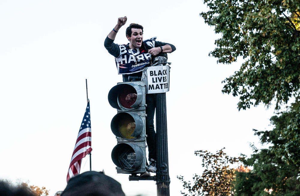 <strong>WASHINGTON, D.C.:</strong> A person from the crowd climbs a traffic stop in front of the White House gates to cheer on the crowd as people clelebrate Joe Biden and Kamala Harris winning the election.