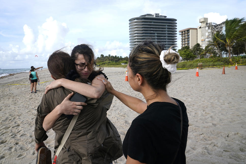 Faydah Bushnaq, of Sterling, Va., center, is hugged by Maria Fernanda Martinez, of Boca Raton, Fla., as they stand outside the partially collapsed building on Friday. Bushnaq is vacationing and stopped to write 