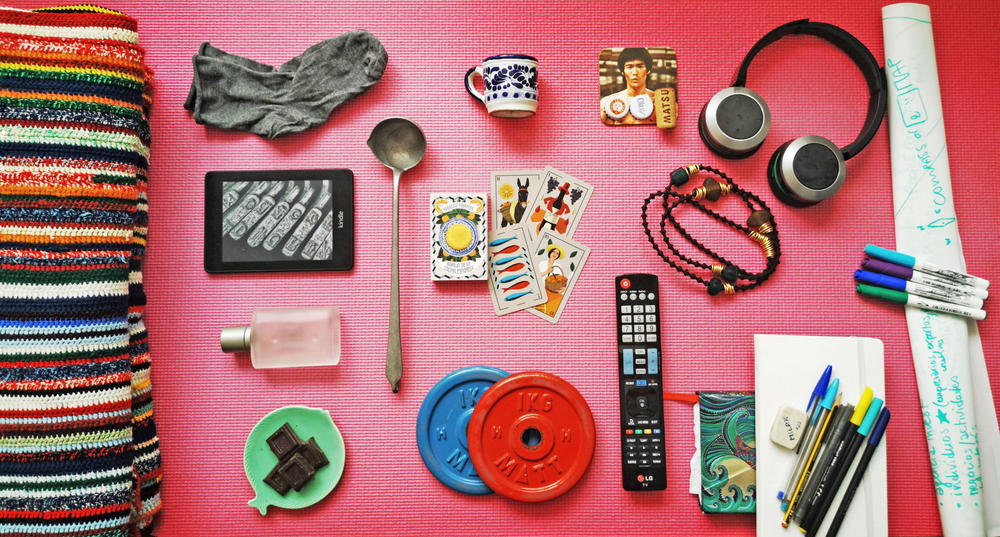Items left to right, top to bottom: Wool quilt, socks, Kindle, perfume, dark chocolate, metal ladle, espresso mug, playing cards, weights, Bruce Lee coaster with bottle caps, necklace, remote, headphones, notebook and pens, whiteboard sheets and markers, all displayed on a yoga mat.