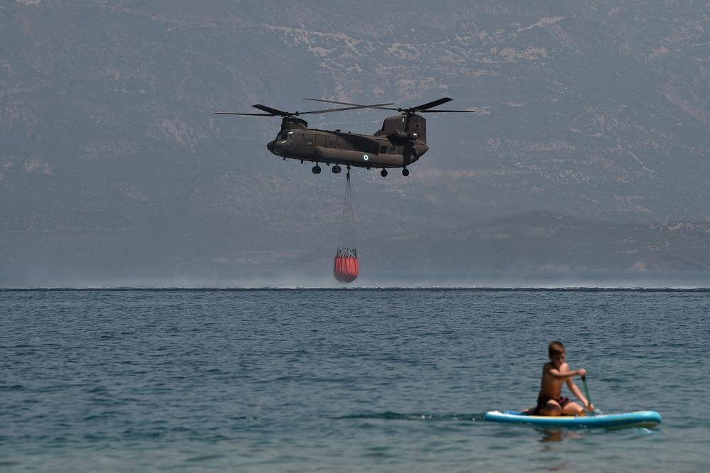 A Ch-47D Chinook helicopter is watched by a paddleboarder as it fills up with water while firefighting near Lambiri Beach at Patras on August 1, 2021. - Nearly 300 firefighters, two water bomber planes and five helicopters were battling to put out a forest fire in Greece that has so far destroyed around 20 homes and injured eight people, authorities said.