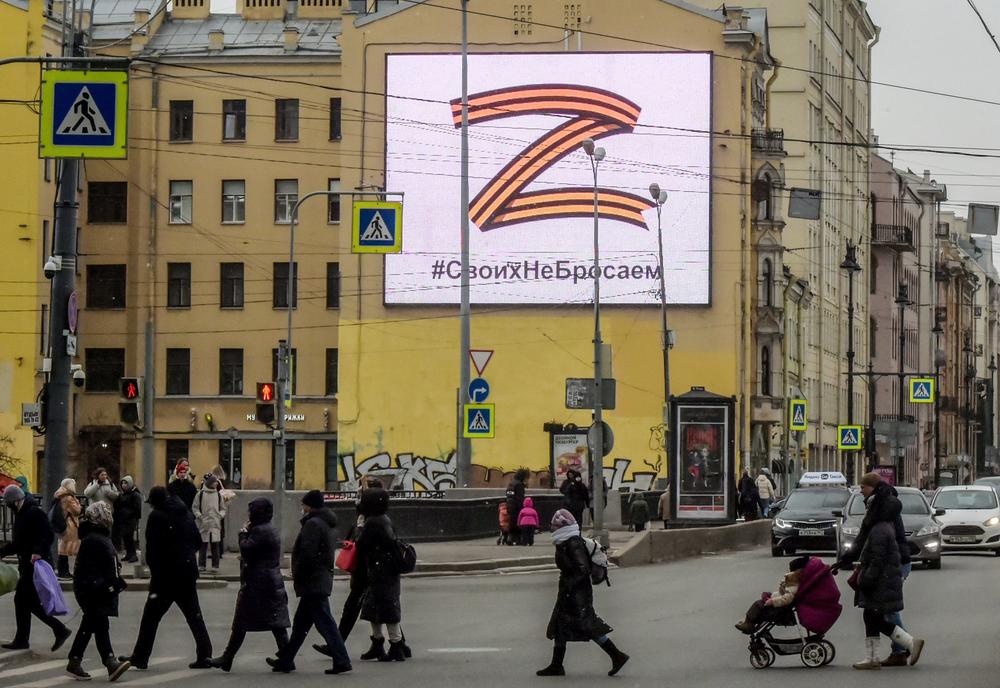 Pedestrians cross a street in front of a billboard displaying the letter 