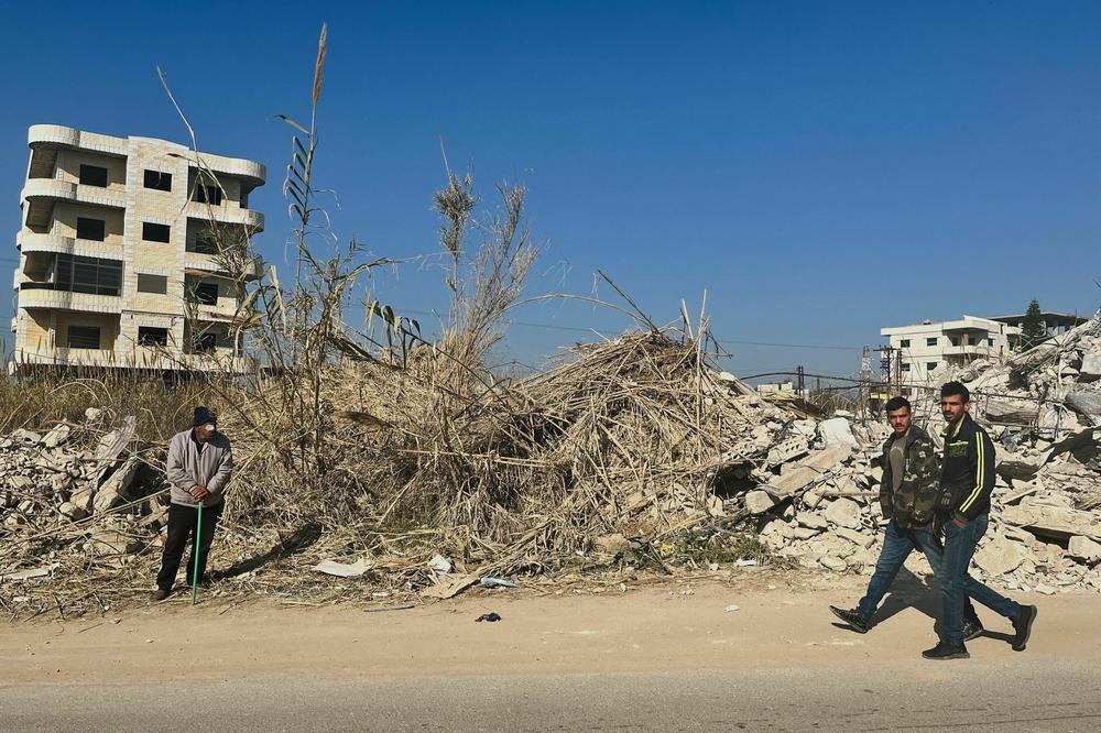 A man stands in front of a home that was destroyed during the earthquake as two men walk by. One of the men is wearing military camouflage. Men in Jableh wear their uniforms long after they've served in the military because it is often the only durable winter wear they have.
