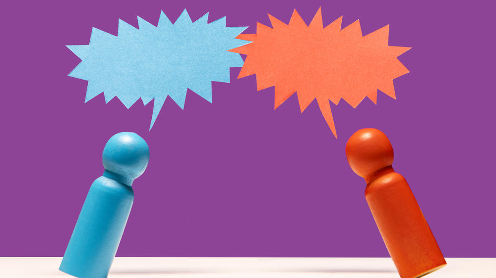 One blue and one red wooden figurine leaning into each other under speech bubbles on white surface, purple background
