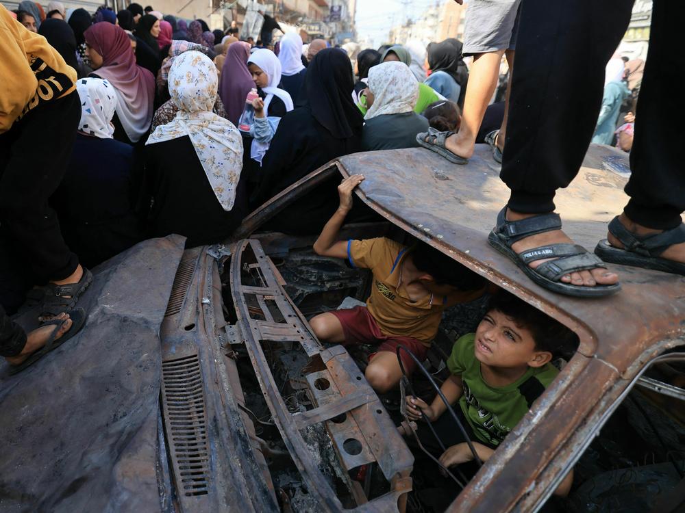Children play in a charred car as people queue for bread in front of a bakery that was partially destroyed in an Israeli strike, in the Nuseirat refugee camp in the central Gaza Strip, on Thursday as battles continue between Israel and the Palestinian Hamas movement.