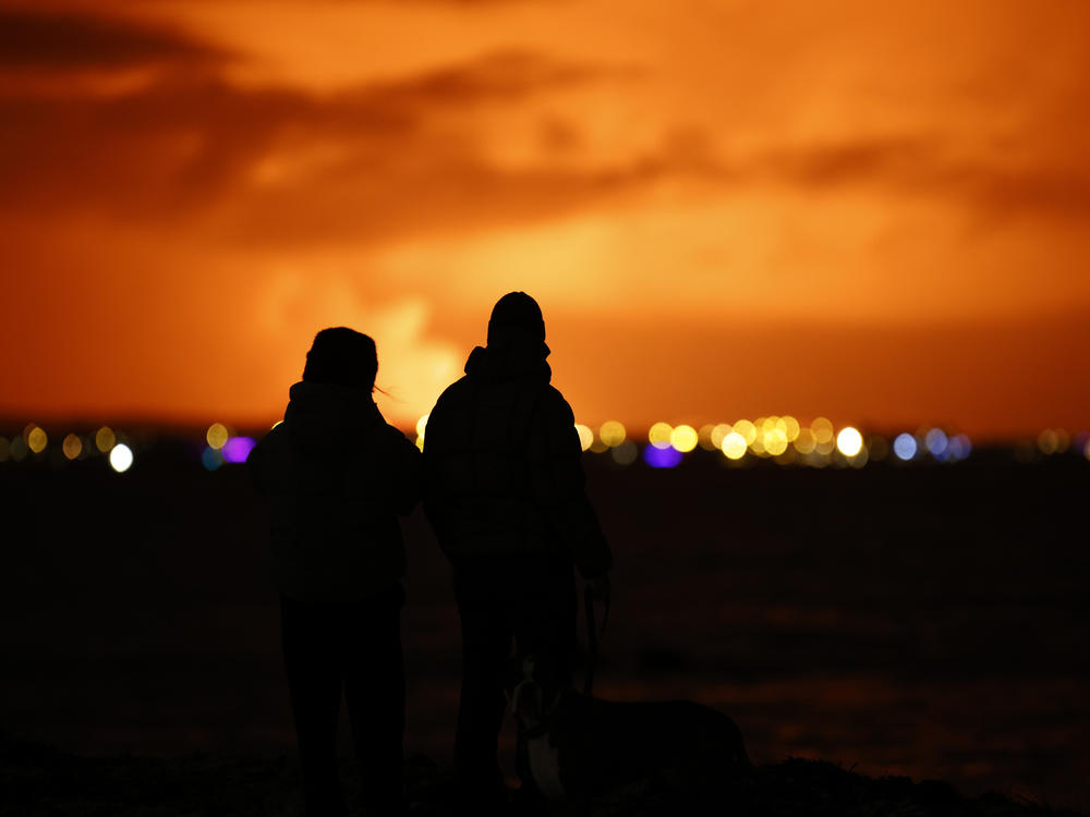 People watch as the night sky is illuminated caused by the eruption of a volcano on the Reykjanes Peninsula in southwestern Iceland seen from the capital city of Reykjavik on Monday, Dec. 18, 2023.