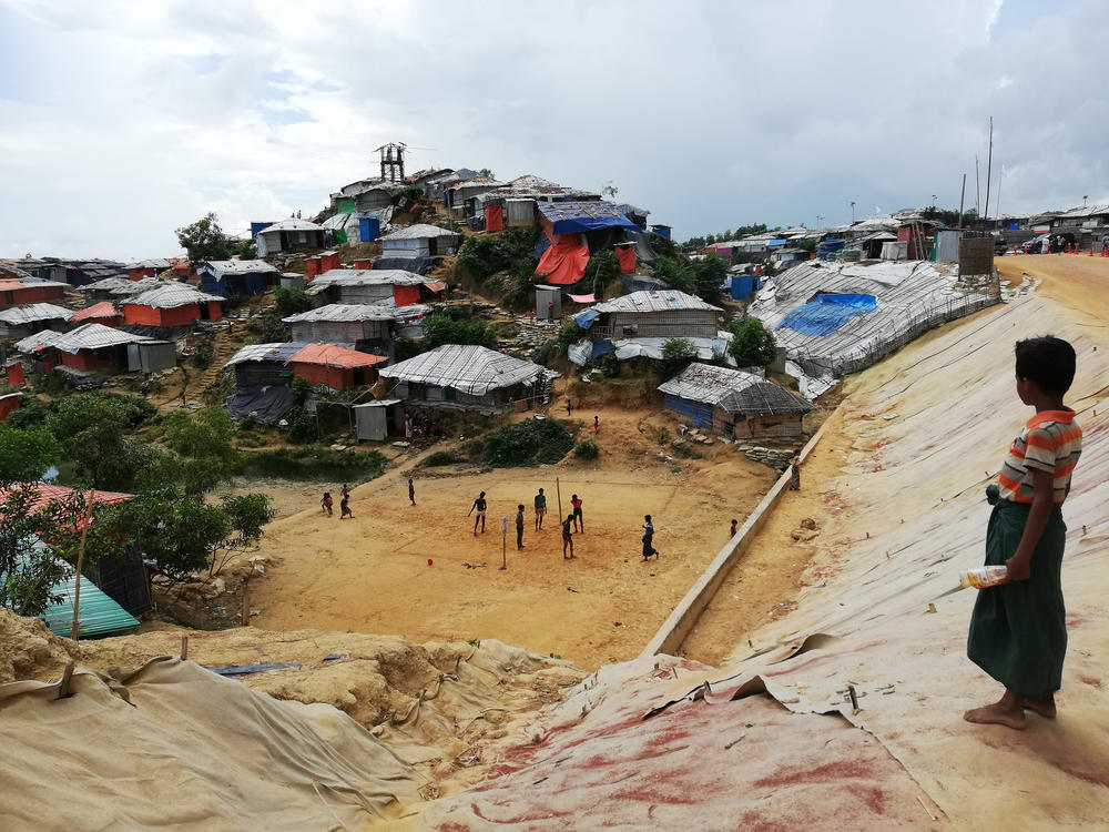 A Rohingya boy watches a game of <em>chinlone</em>, also known as caneball, a traditional sport from Myanmar. The rooftops of refugee homes in the distance are typically made of bamboo and tarpaulin.