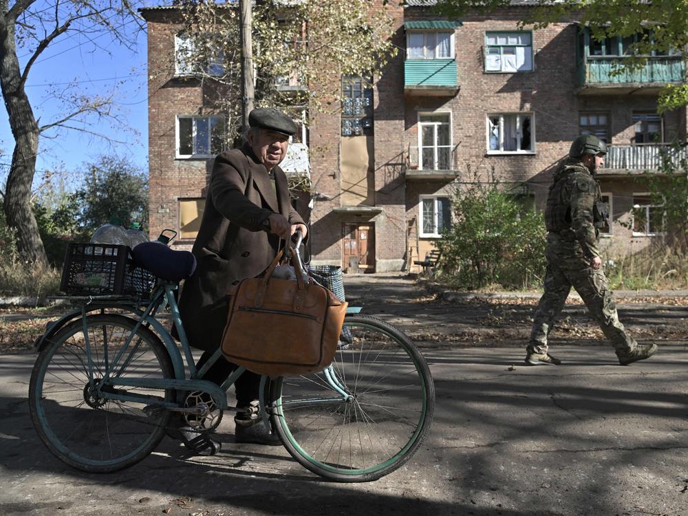 A Ukrainian policeman (R) walks next to 84-year-old resident Mykola (L) pushing his bicycle on a street in the frontline town of Chasiv Yar, in the Donetsk region on October 11, 2023, amid the Russian invasion of Ukraine. Explosions are near-constant in the small town, whose buildings are scored with holes from shelling.
