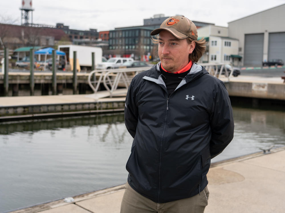 Portrait of Alex Snider wearing his Oriole hat on opening day, standing on a pier at a Baltimore marina and talking about the impact the bridge collapse will have.