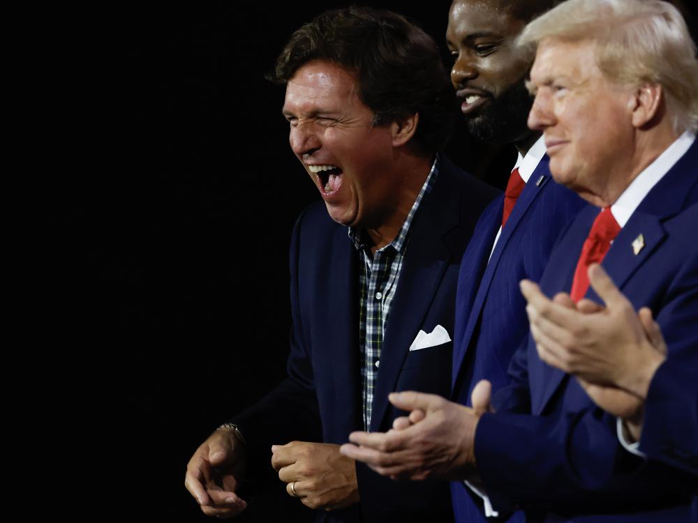 Right-wing personality Tucker Carlson laughs as he stands alongside Congressman Byron Donalds, Republican presidential candidate Donald Trump, and vice presidential candidate J.D. Vance on the first day of the Republican National Convention in Milwaukee.