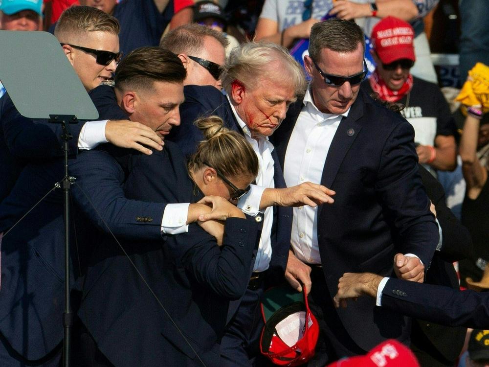Blood is on Trump's face and right ear as Secret Service agents surround him and take him offstage amid the assassination attempt.