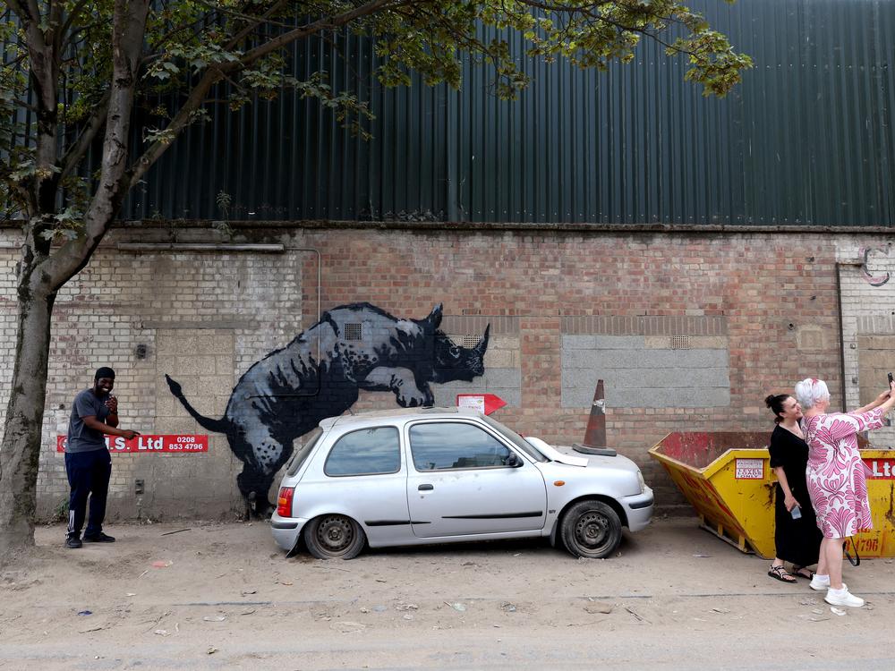 People gather around an artwork by street artist Banksy, the eighth to released in eight days, depicting a rhino mounting a car, on a wall in Charlton, London, on Aug. 12.