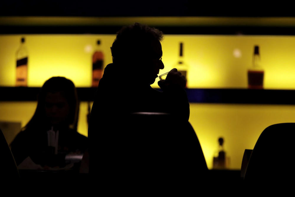 A person sits at a bar with a hand holding a glass to the mouth 