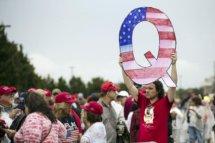 A man in a red shirt holding up a large "Q" shaped sign in a protest. The "Q" is colored in with the shapes and colors of the American flag. Other protesters around him wear red MAGA hats.