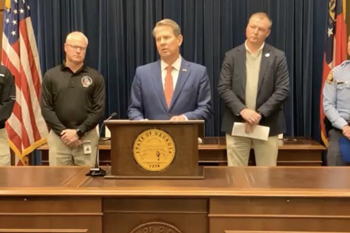 Governor Brian Kemp is shown standing behind a desk and lectern flanked by state officials.