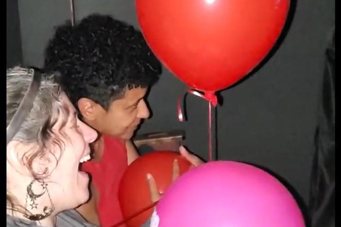 Two red balloons float in the air as two people laugh below while holding balloons