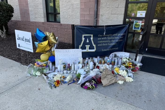 Flowers, balloons, signs, candles and other symbols of support are shown on a sidewalk next to a door.