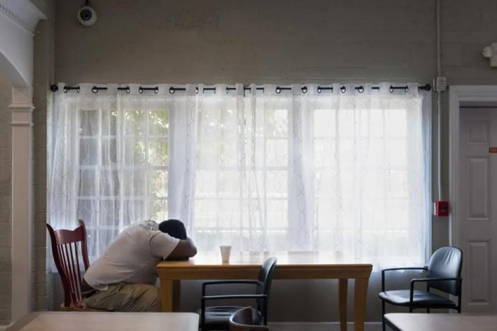 A man is shown sitting on a chair with his head facing down onto his arms resting on a table inside an empty room.