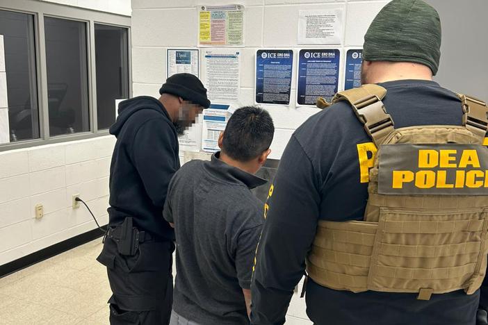 Three individuals are shown from behind, standing in a drab room.  One of them wears a uniform that says "DEA Police."